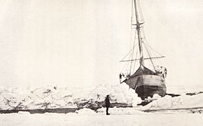  Vue de face d'un navire penchant vers tribord. Il est entouré de glace mais, devant la proue, une étroite nappe d'eau est visible. La scène est observée par un homme débout sur la glace.