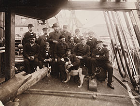  Groupe de 13 hommes et un chien posant sur le pont étroit d'un navire, au milieu des cordes, des épars et du gréement, tous vêtus de chapeau et, à une exception près, de costumes sombres.