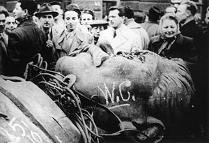  A group of 17 civilians behind the head of a fallen bronze statue of Josef Stalin lying face-up, defaced with some graffiti including "W.C."