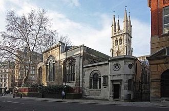 À la droite de la photo apparaît la façade d'un immeuble. La face avant comprend plusieurs fenêtres en ogive. Un arbre est à la gauche de l'église. À l'arrière de celle-ci, une tour de style gothique la surplombe.