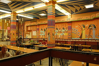 Grande salle compartimentée, comportant table et chaises, et dont les murs sont décorés de motifs (personnages, frise d’oiseaux, dessins géométriques) inspirés par les fresques égyptiennes antiques. La couleur orange domine l’ensemble.
