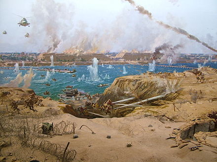 Peinture montrant les hommes du Génie égyptien utilisant les pompes pour détruire la barrière de sable de la ligne Bar-Lev, avec en fond les bateaux, les hélicoptères traversant le canal tout cela sous le feu incessant de l'artillerie.